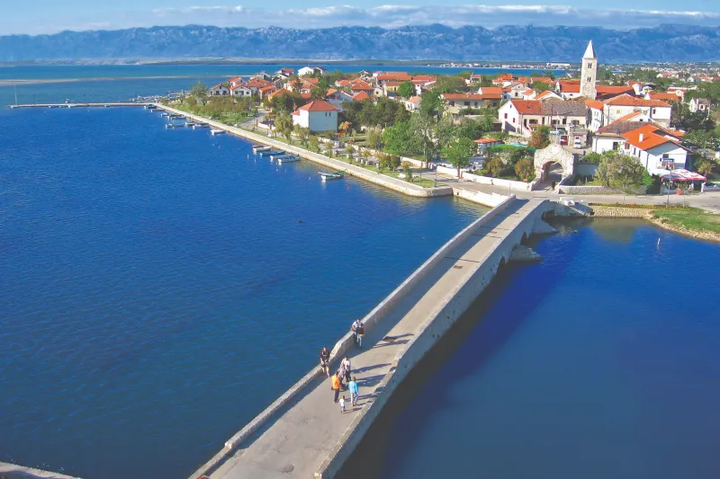 Apartments Nin – Aerial view of the stone bridge leading to the historic town of Nin, surrounded by blue sea and red rooftops along the bay.