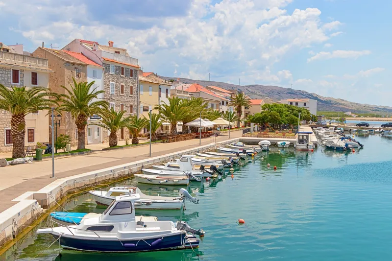 Apartmani Pag – Obalna šetnica Paga sa starim kamenim kućama, palmama i vezanim brodicama u čistom plavom moru, s blagim planinama u pozadini.