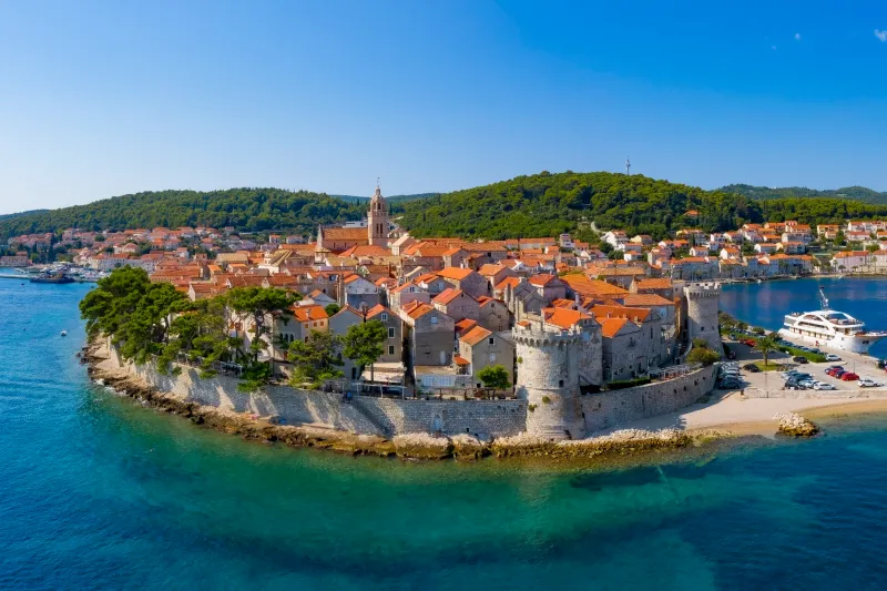 Korčula appartamenti – vista panoramica sulle mura e torri della città vecchia di Korčula, mare turchese, pini e barche a vela in una giornata di sole.