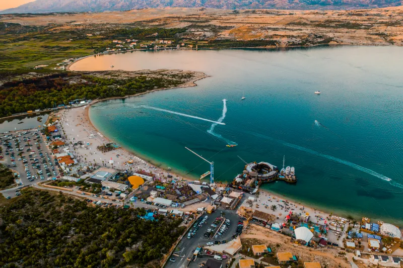 Novalja apartamentos – vista aérea de la playa Zrće con mar cristalino, clubes, barcos y muchos visitantes en un día soleado en la isla de Pag.