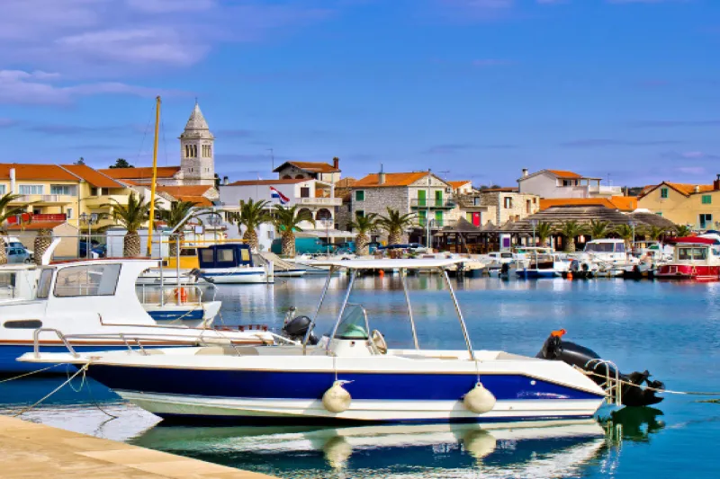 Pier and boats in Pakoštane, a picturesque Dalmatian coastal town, with palm trees, an old church and traditional houses in the background.