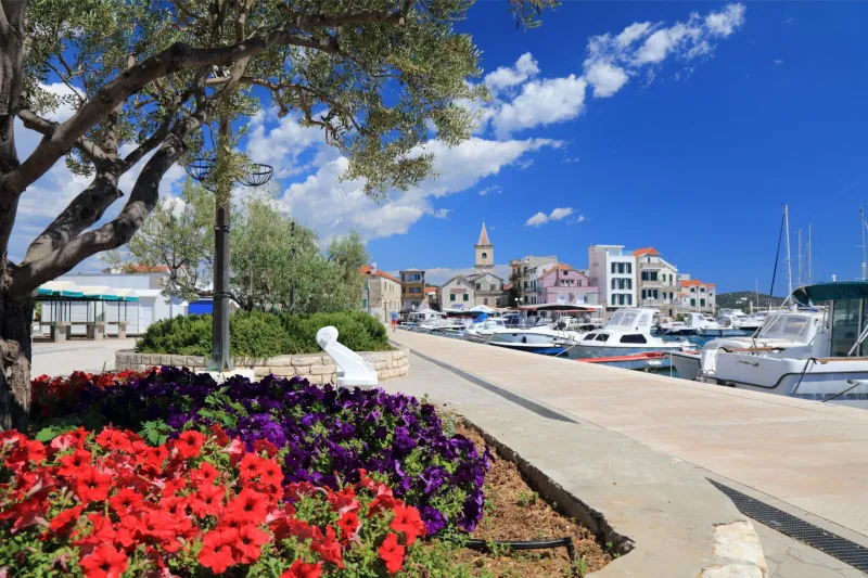 Pirovac appartamenti – vista sul lungomare mediterraneo di Pirovac con aiuole fiorite, alberi, barche in marina e case colorate sotto il cielo blu.