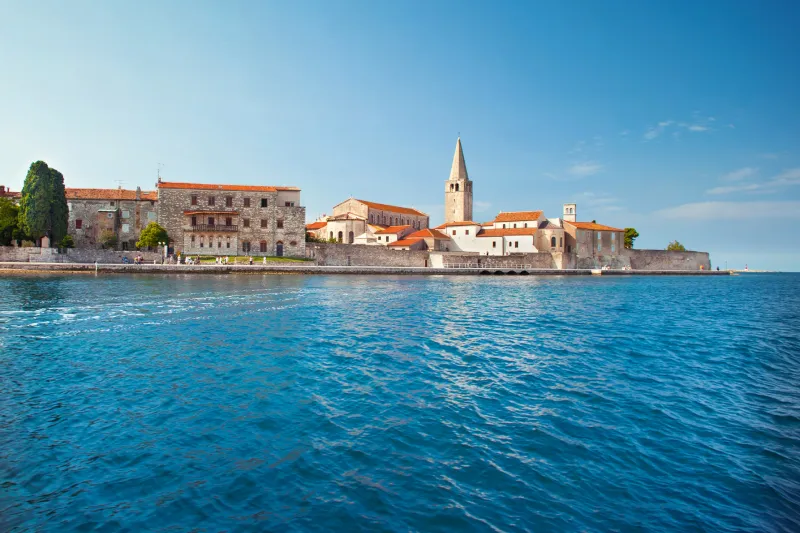 Poreč apartmani – Pogled na stari grad Poreč s mora, s prepoznatljivom bazilikom i tornjem, kamenim zgradama i plavim Jadranskim morem pod vedrim nebom.