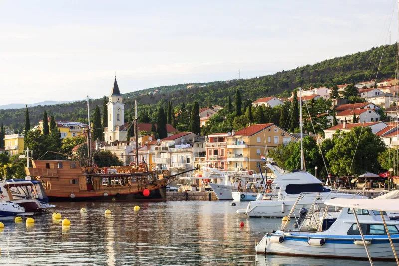 Selce apartments - picturesque view of the marina with a wooden boat, white yachts and colorful houses along the green hillside.